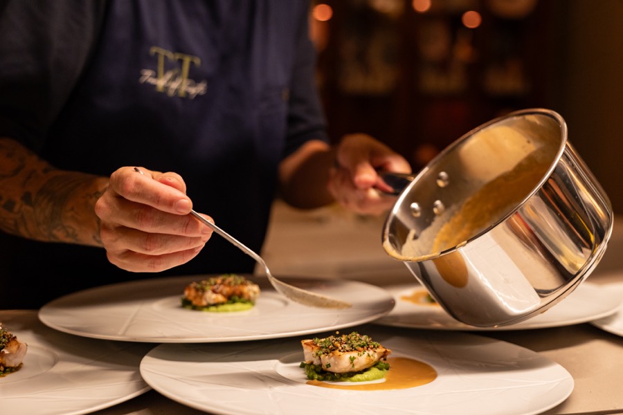 Chef spooning sauce onto plated fish from a small saucepan